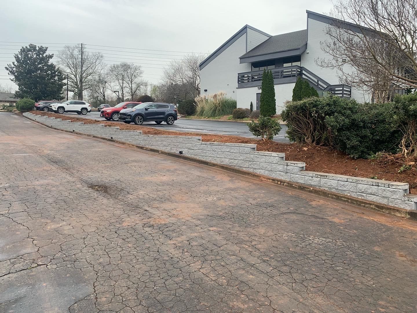 Asphalt parking lot with cars, low brick retaining wall, and two-story white building. Overcast sky.
