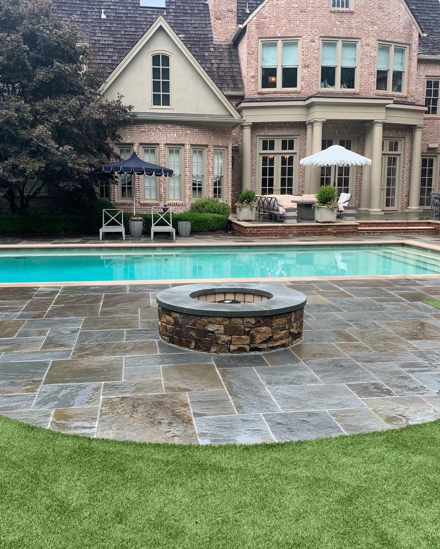 Backyard pool and patio with a fire pit, overlooking a large brick house.
