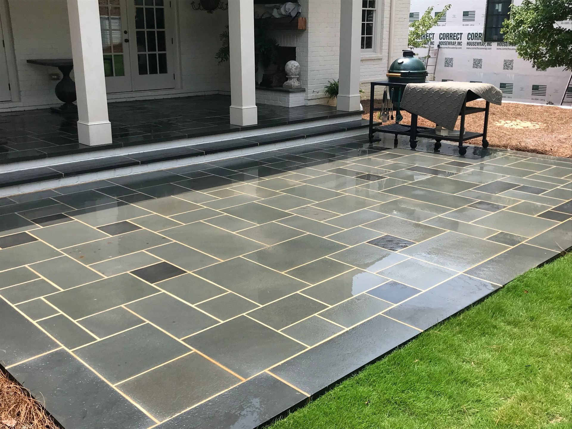 Patio with blue-gray stone tiles, a black grill, and green grass.