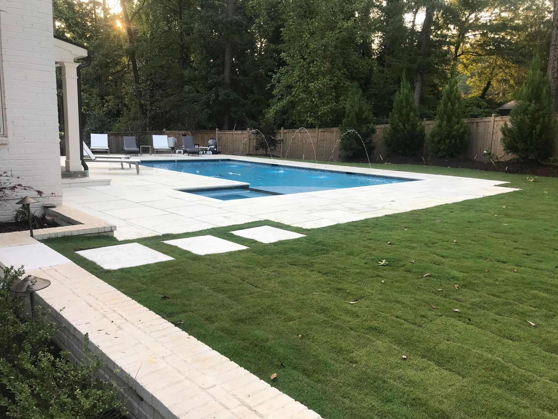 Backyard with a swimming pool, spa, light-colored patio, green grass, and trees.