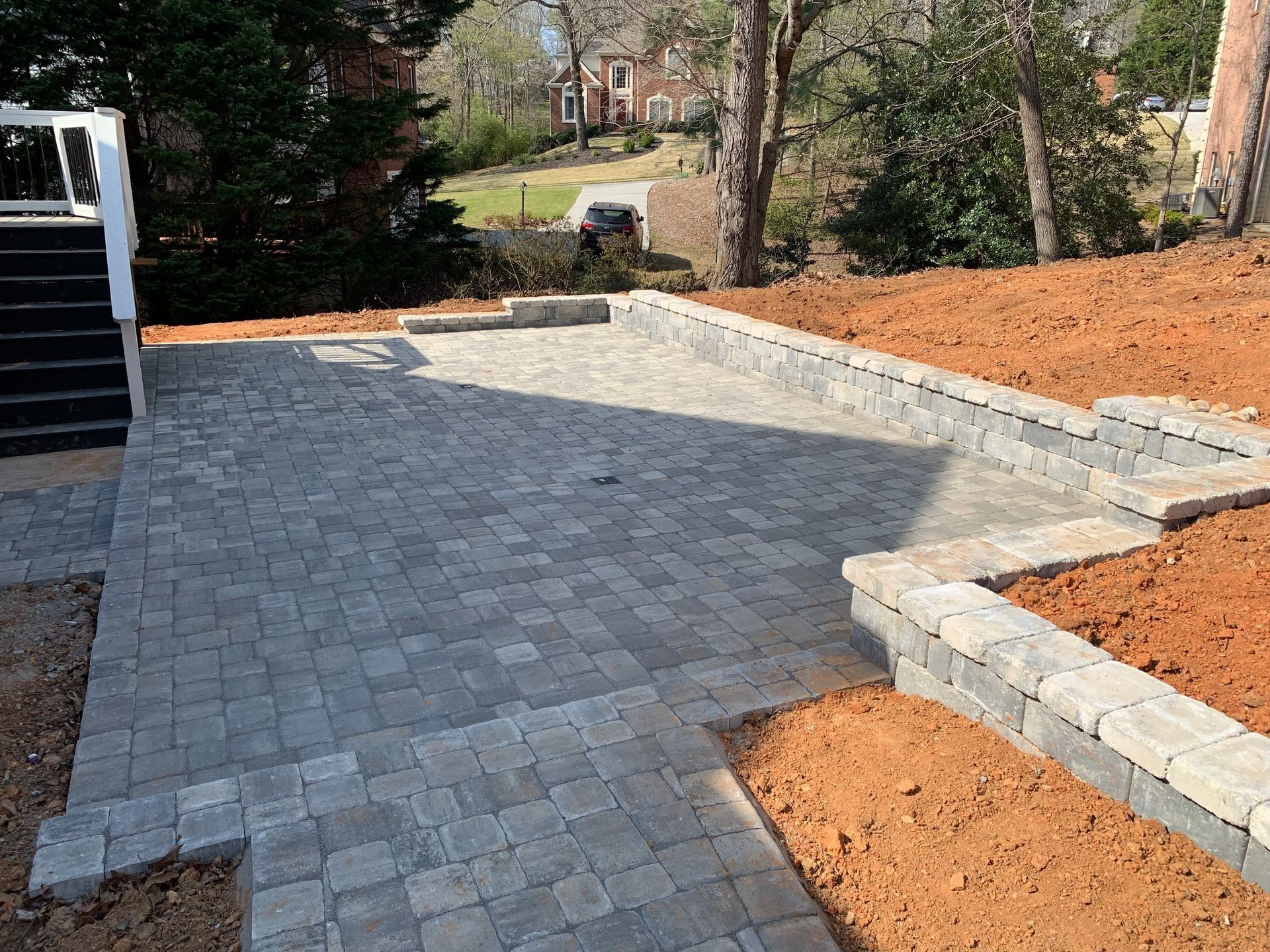 Brick patio construction with retaining walls and steps leading to a yard with bare soil and trees.