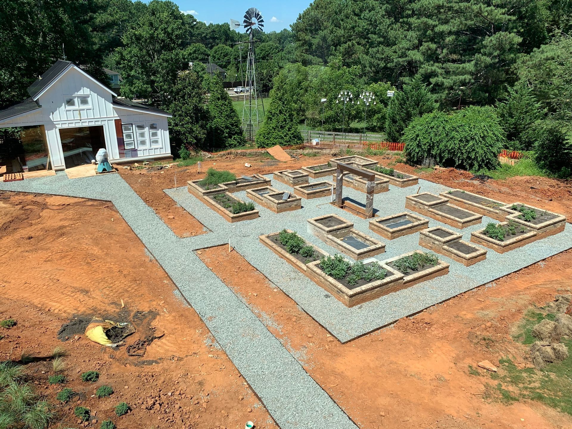 A raised garden bed layout with gravel paths, a white shed, and a windmill in a rural setting.