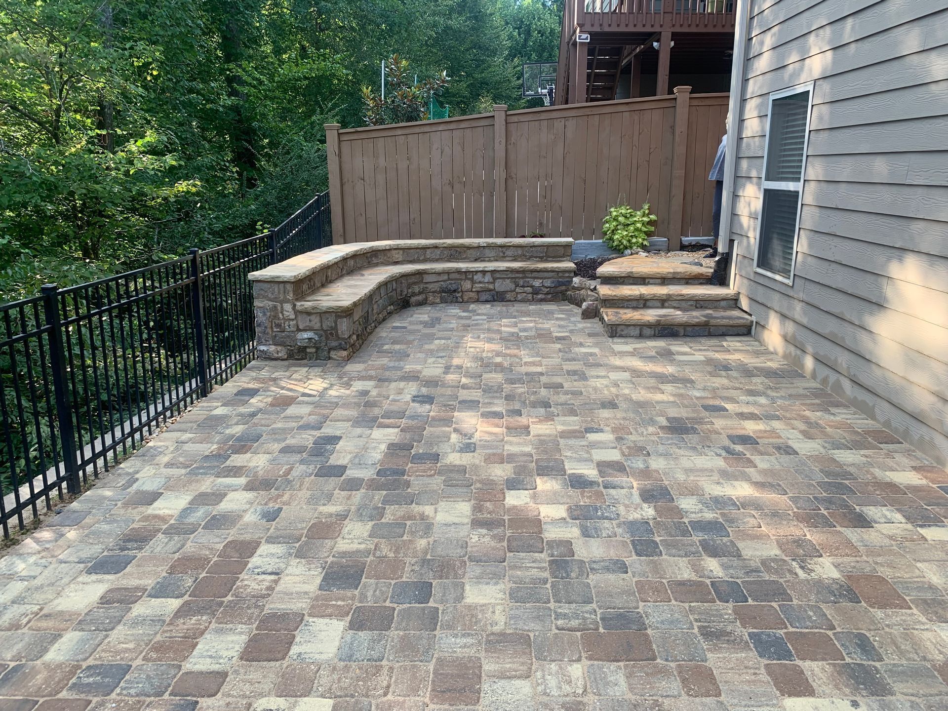 Brick patio with stone bench, steps, and black metal railing near a house and trees.