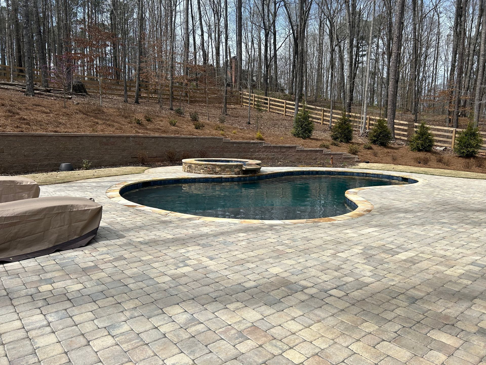 A dark blue kidney-shaped pool with stone patio. Trees and a wooden fence are in the background.