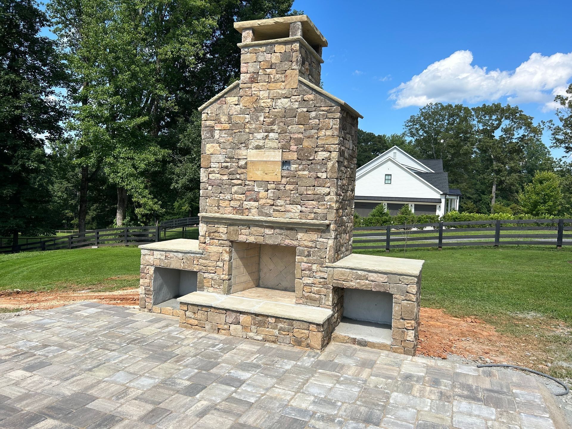Stone outdoor fireplace with side storage, on a paved patio. Trees and a house in the background.