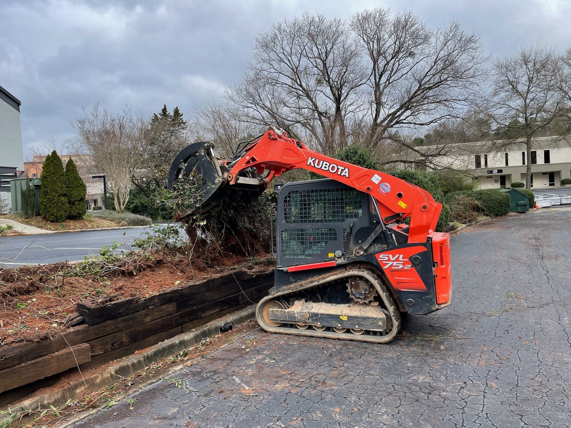 Orange Kubota skid steer clearing bushes on a paved area next to a building.