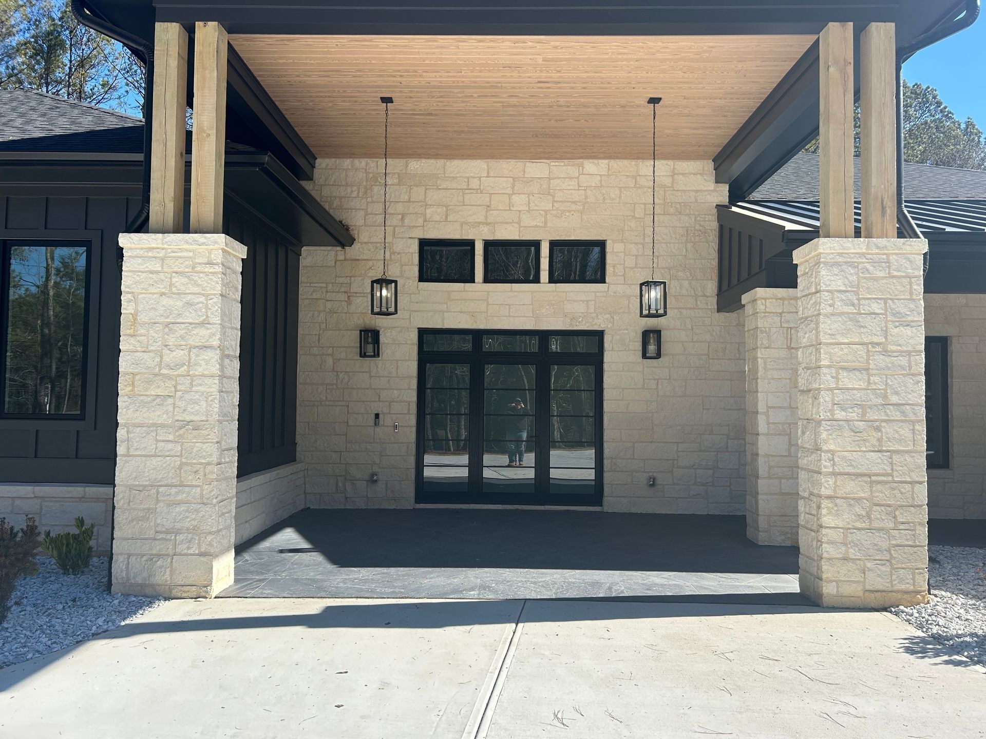 Stone-faced entry of a modern building with a black framed door and pillars, under a wood-paneled ceiling.