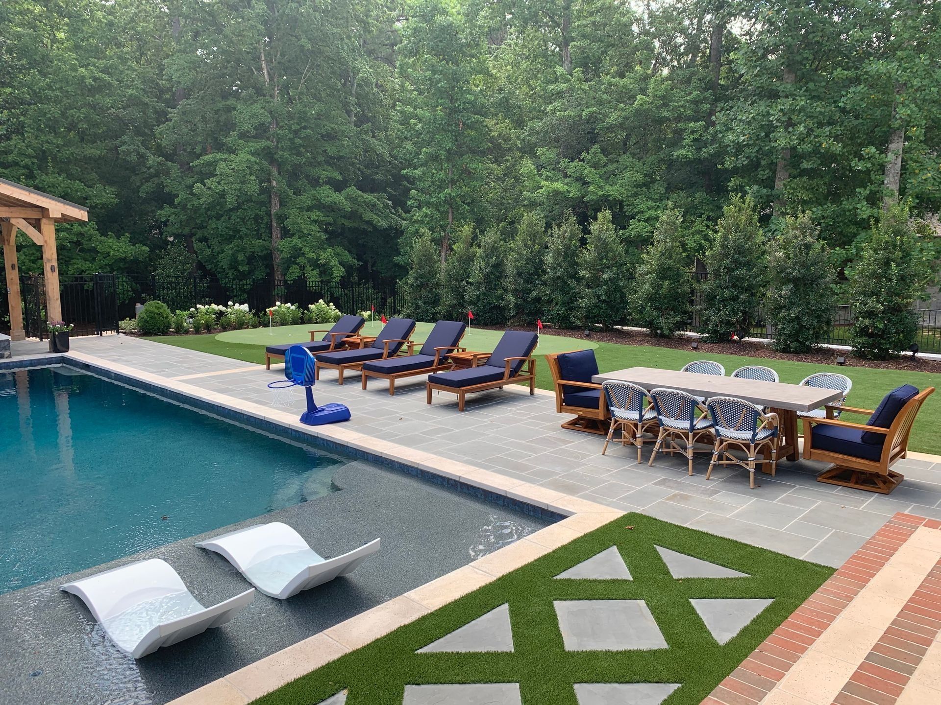 Poolside patio with pool, lounge chairs, dining table, and green lawn with trees in the background.