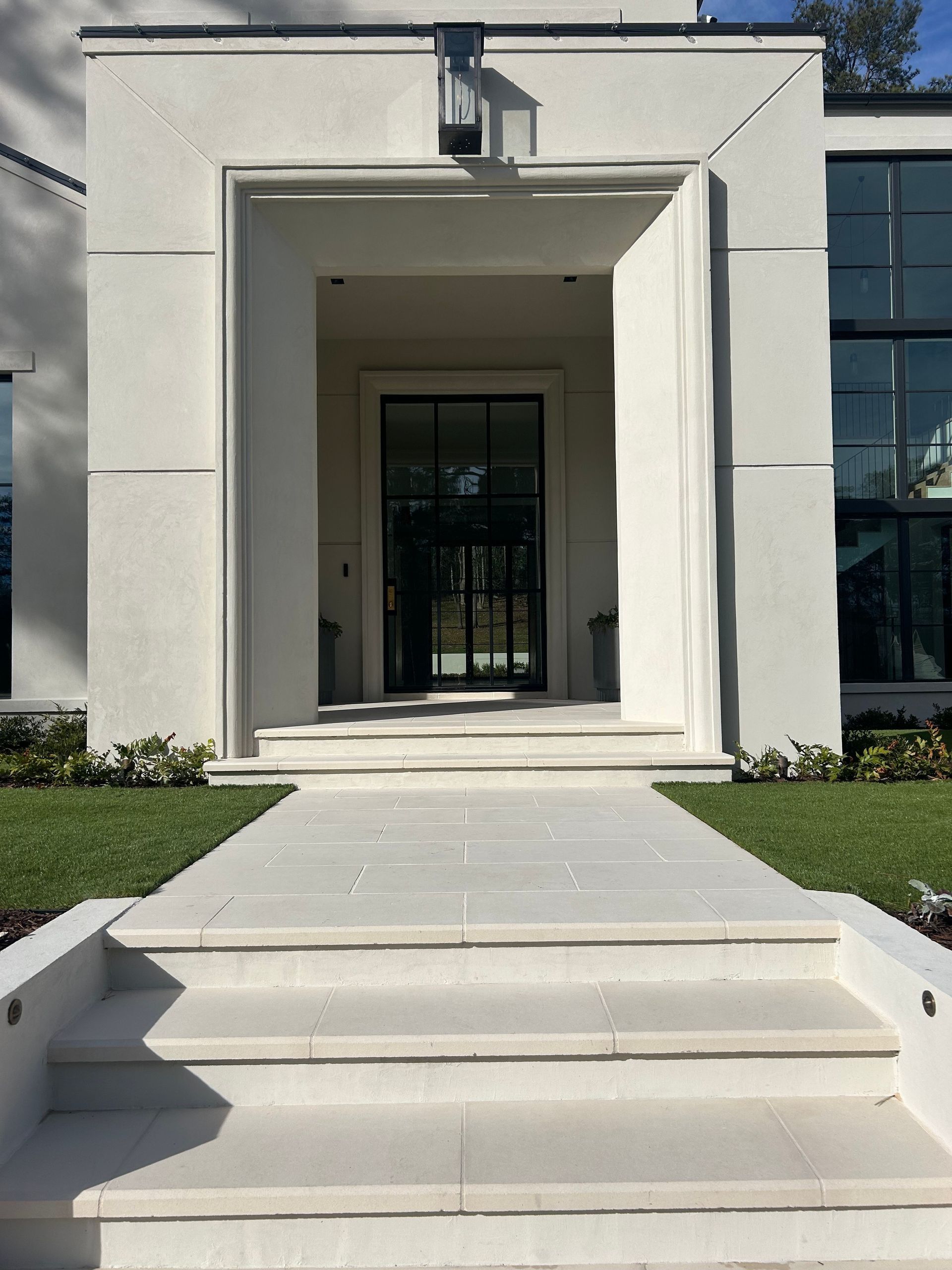 White stone steps lead to a recessed doorway with a black framed glass door, set in a white stucco facade.