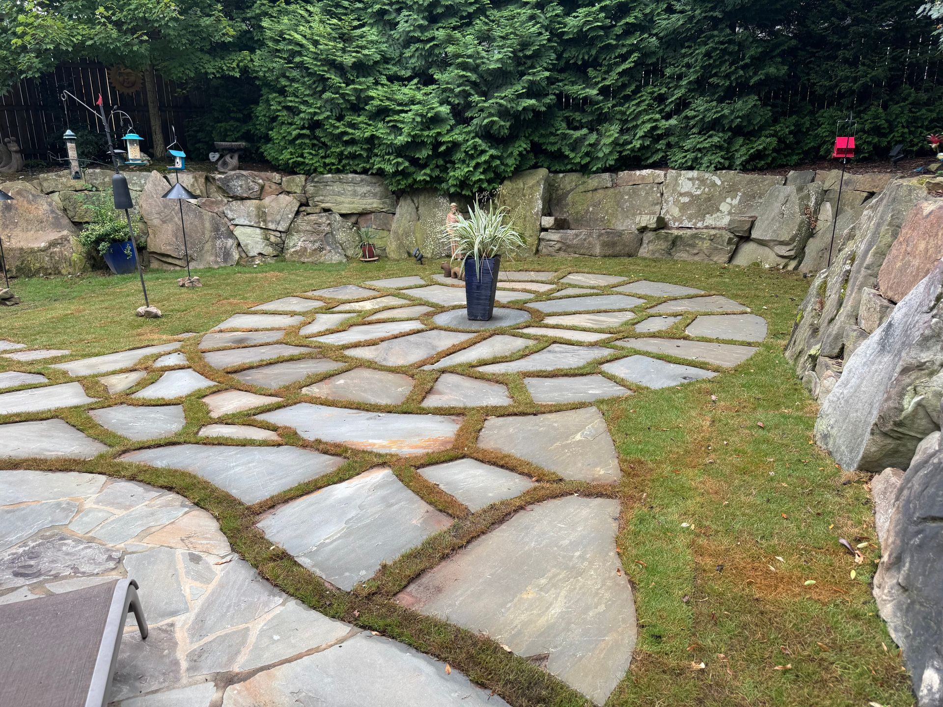 Stone patio with plant-filled gaps, surrounded by grass and rock walls. A vase sits in the center.