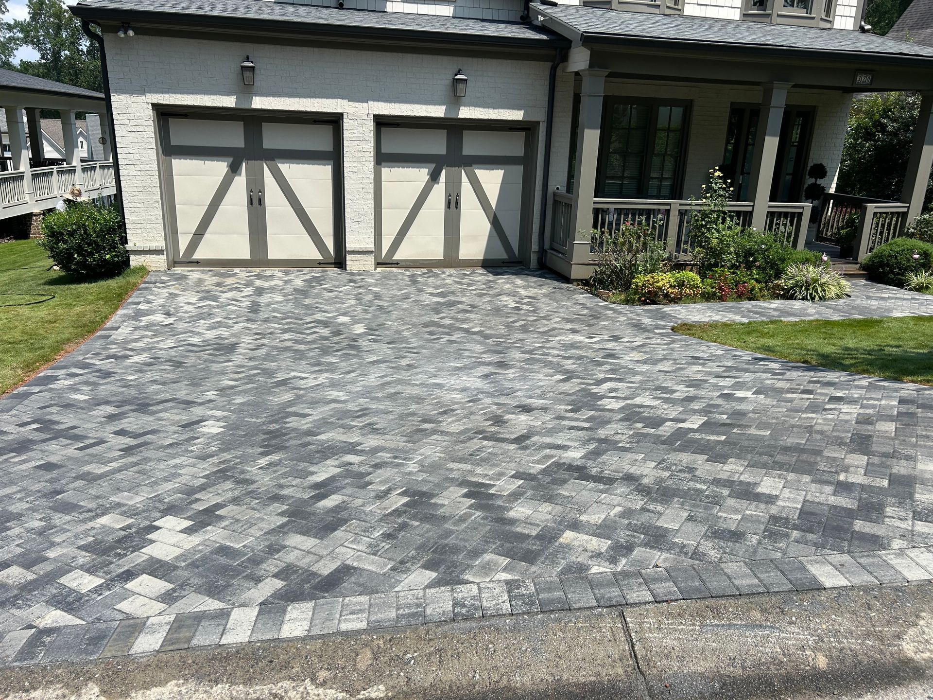 Driveway paved with gray and black bricks leading to a house with a garage and porch.