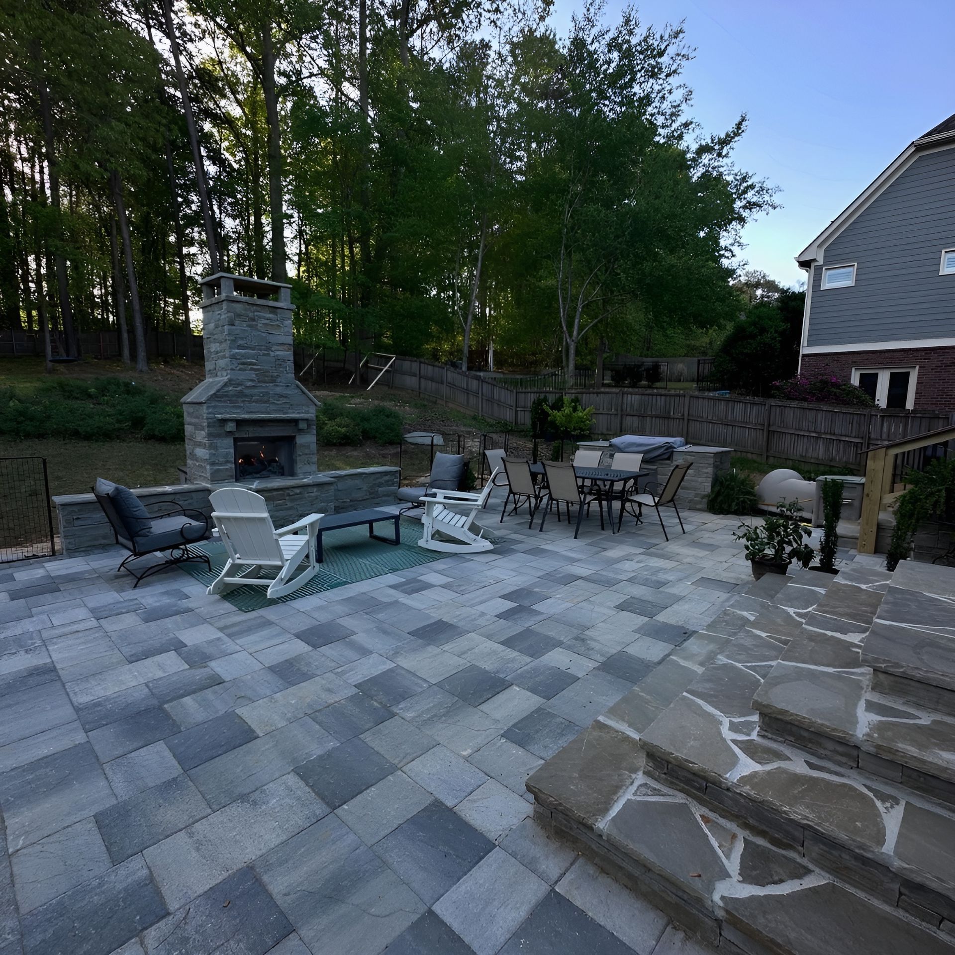 Outdoor patio with stone fireplace, seating area, and pavers, near a house and trees.