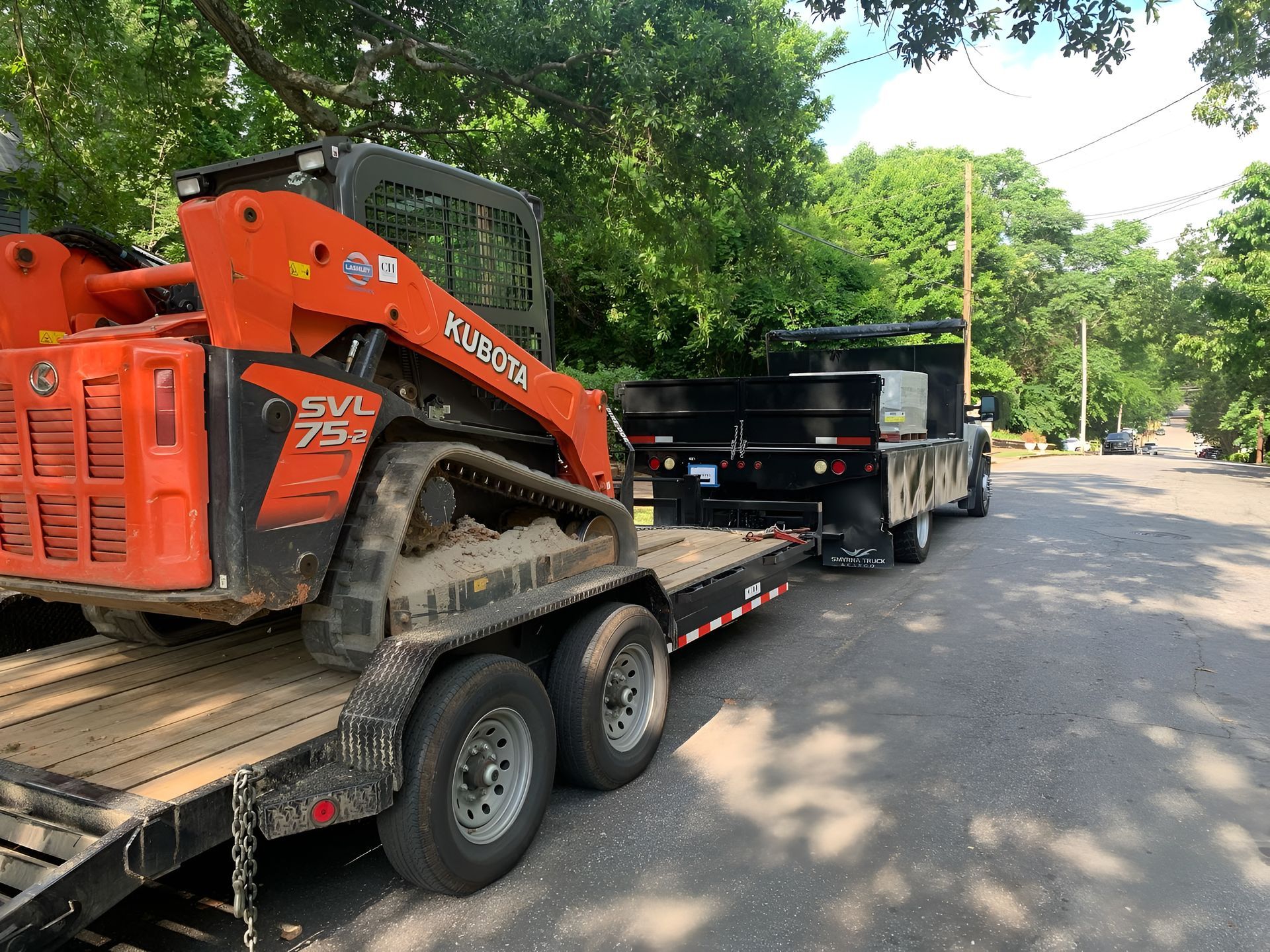 Orange skid steer loader on a trailer hooked to a truck, parked on a road.