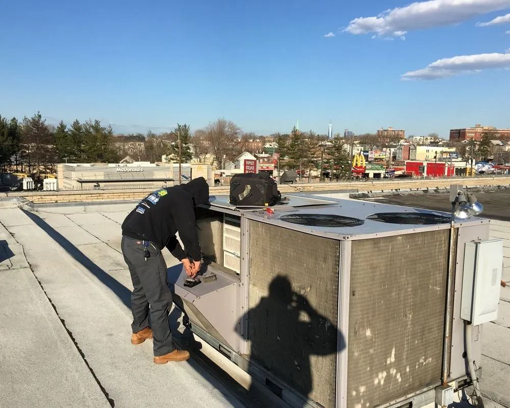 Person in hooded sweatshirt repairs rooftop air conditioning unit. Bright sky and urban background.