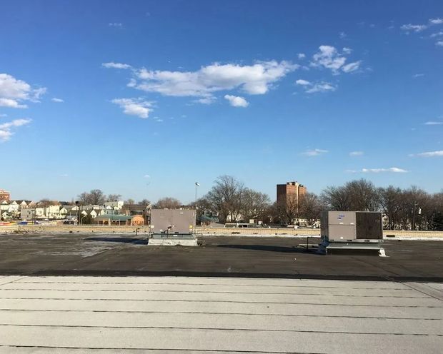 Rooftop with air conditioning units against a blue sky with scattered clouds. Buildings and trees in the distance.