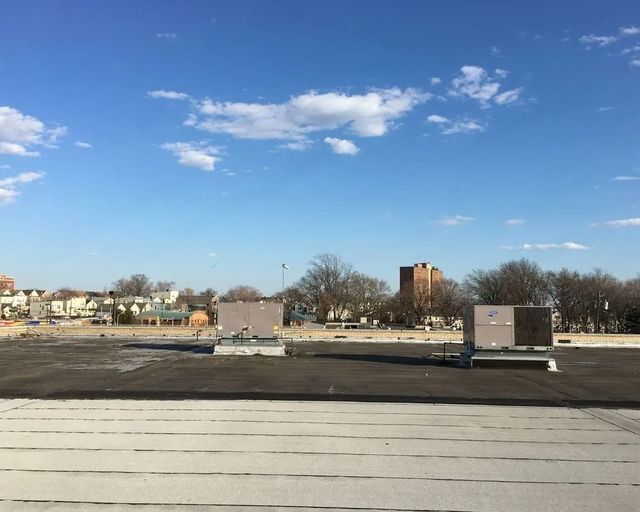 Rooftop with air conditioning units against a blue sky with scattered clouds. Buildings and trees in the distance.