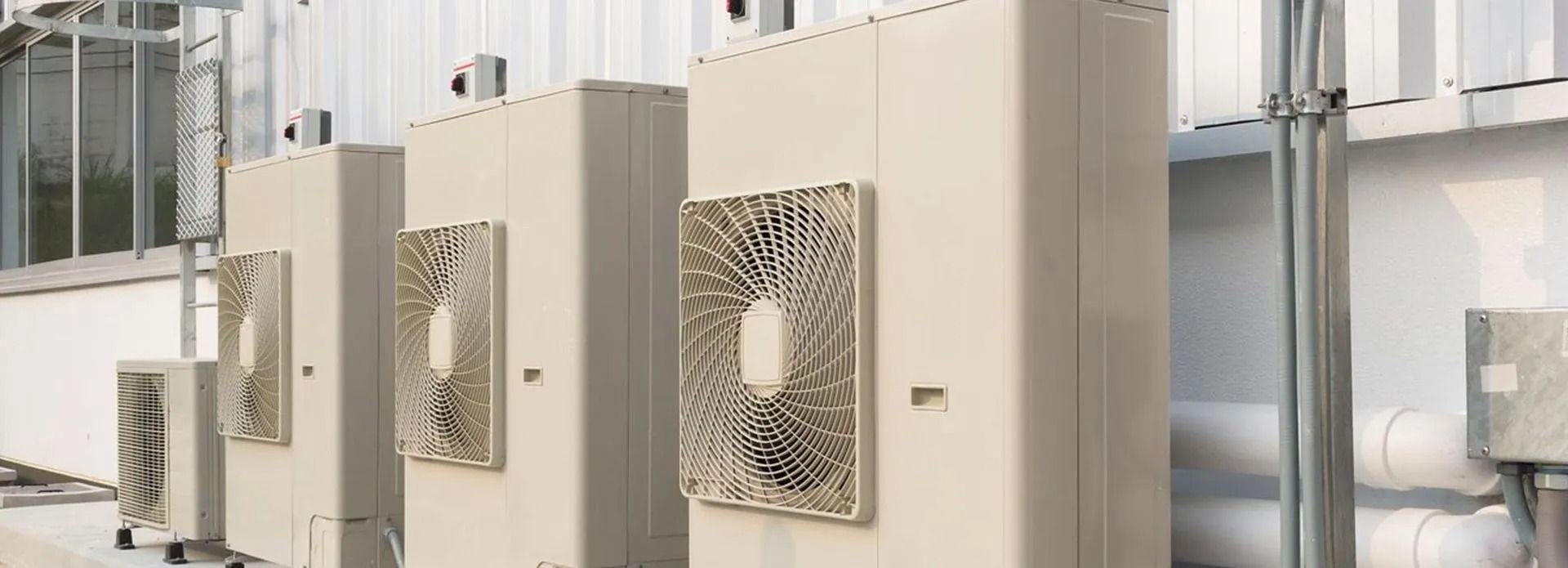 Several large air conditioning units in a row, with exterior fan covers, mounted against a light-colored building wall.
