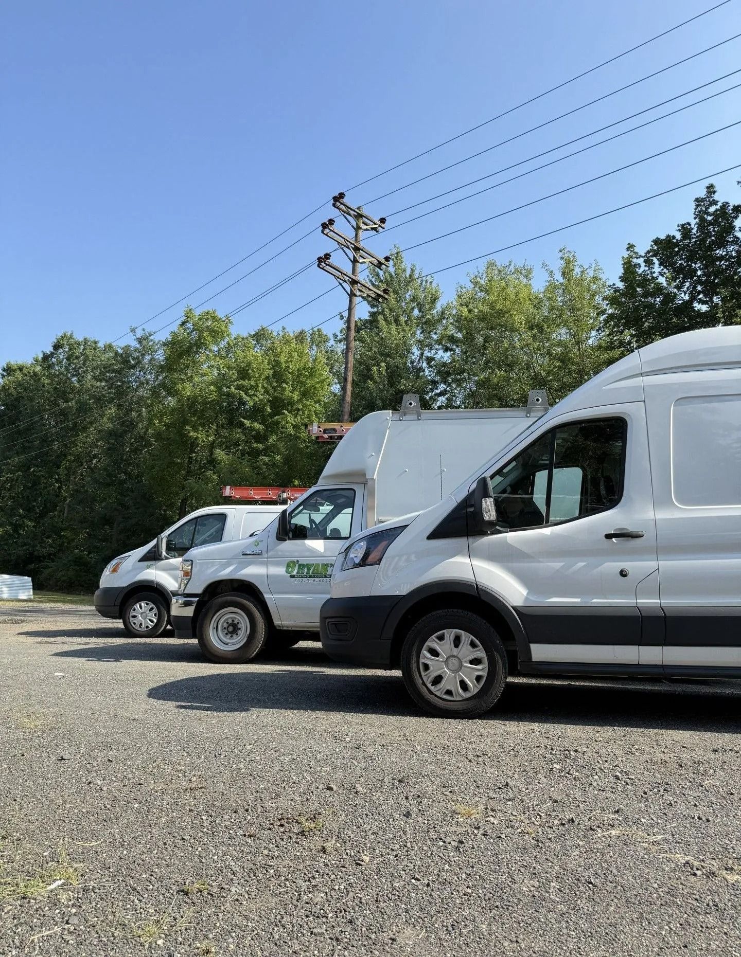 Three white work vans parked on gravel, with trees and power lines in the background under a blue sky.