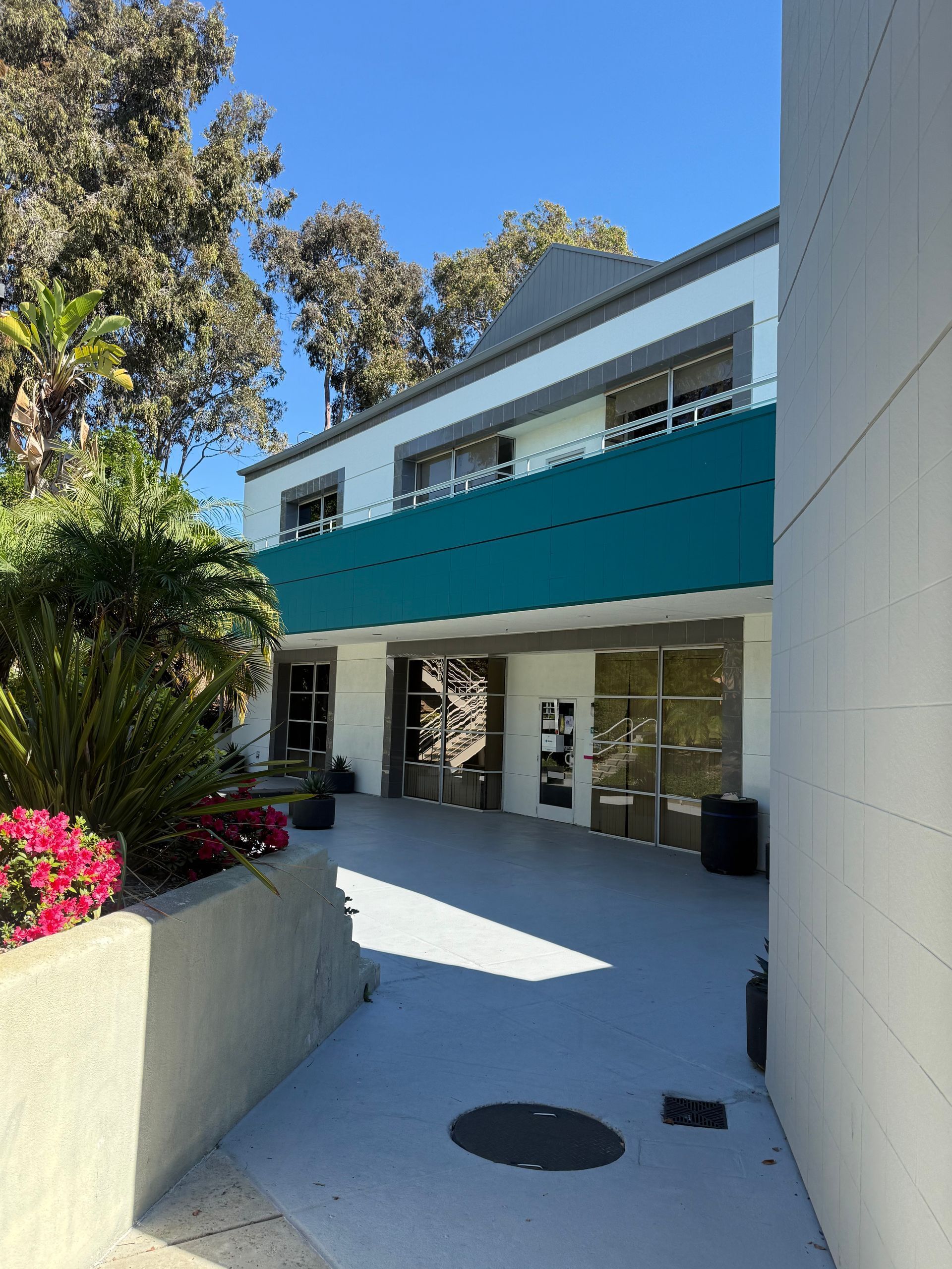 Exterior of a two-story building with teal awning, concrete patio, trees and flowers on a sunny day.