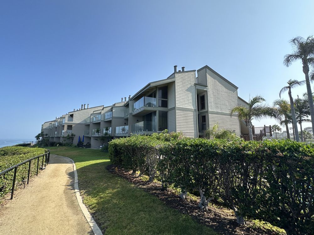 Apartment building overlooking the ocean with a walking path and palm trees under a clear blue sky.