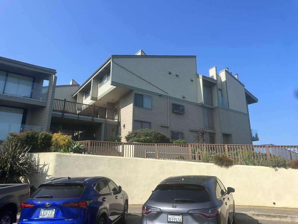 Gray building with multiple levels, balconies, and cars parked in front under a blue sky.