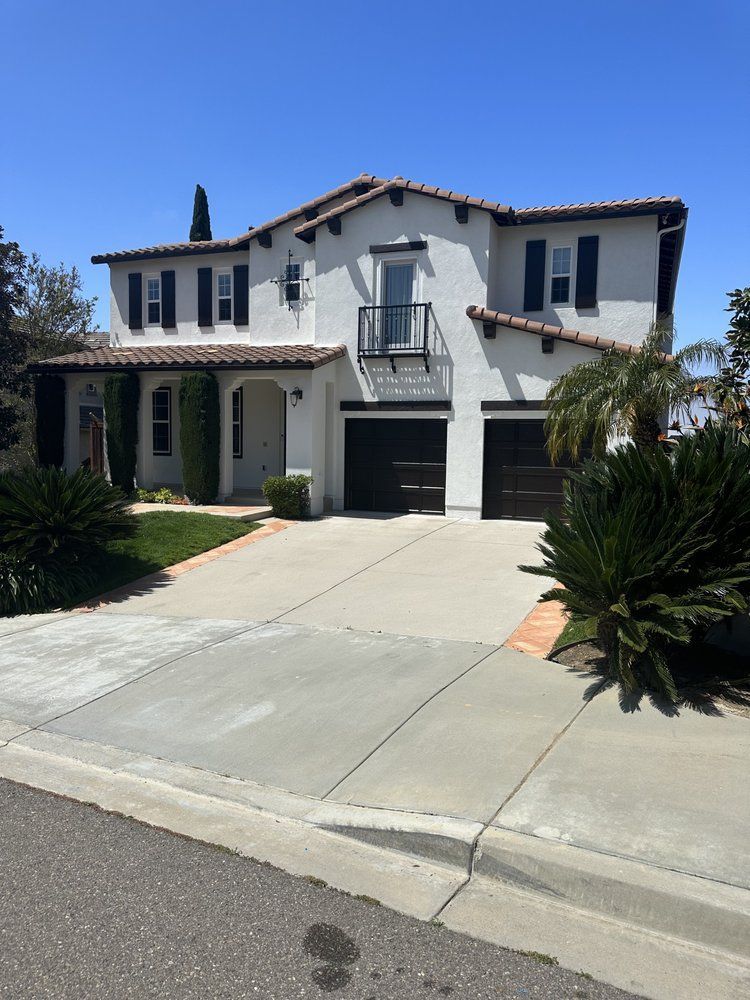 Two-story white house with black shutters and garage doors; driveway; front yard with shrubs; clear blue sky.