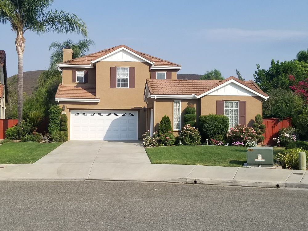 Two-story tan house with white garage door, red tile roof, green lawn, and a clear blue sky.