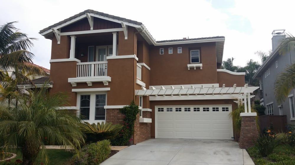 Two-story brown house with white trim, pergola over garage, and palm trees in front.