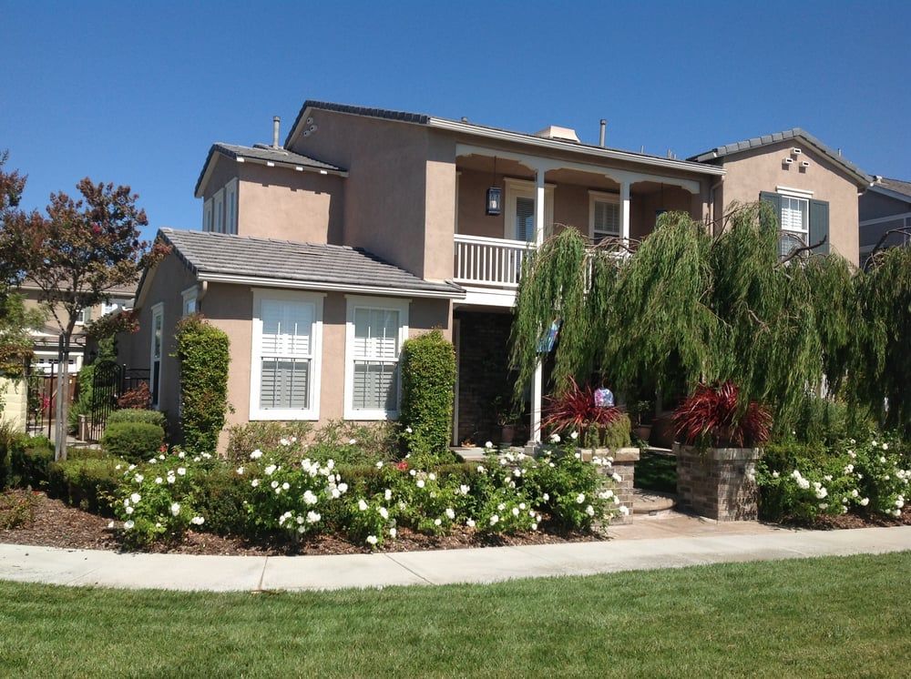 Two-story stucco house with porch and landscaping, surrounded by green grass, under a clear blue sky.