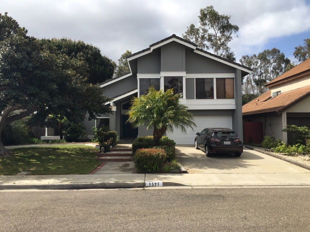 Gray two-story house with a white garage, driveway, and a black car, surrounded by trees and green grass.