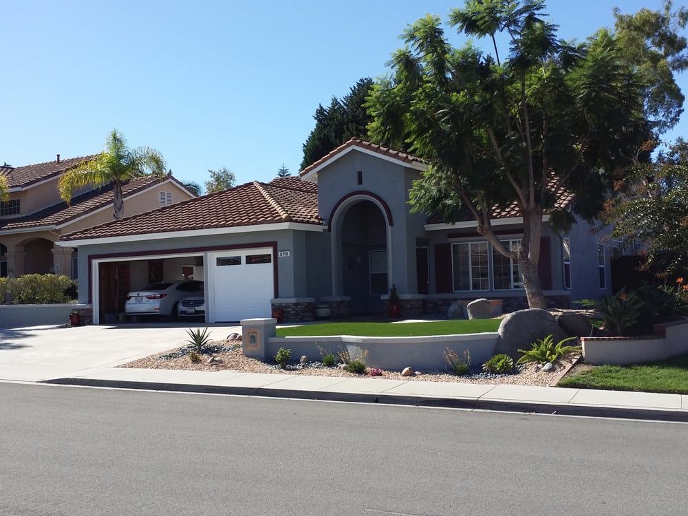 Blue house with red-tiled roof, open garage, and front yard landscaping under a sunny sky.