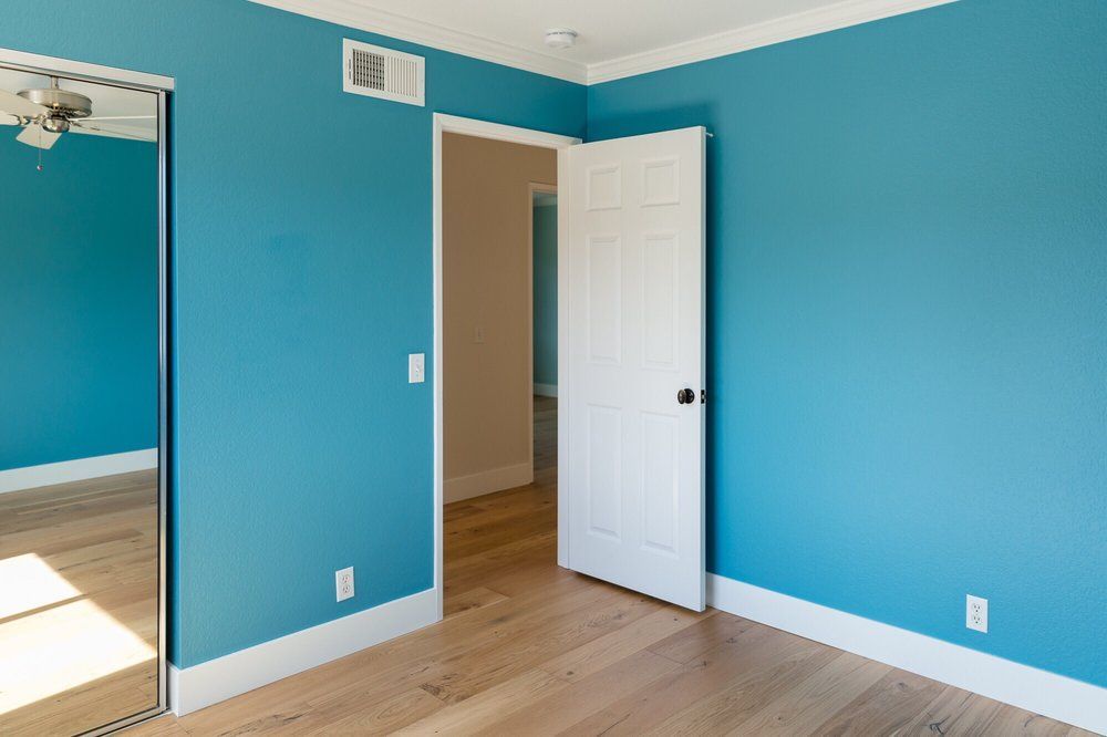 A blue-walled room with an open white door, wood floors, and a large mirror.