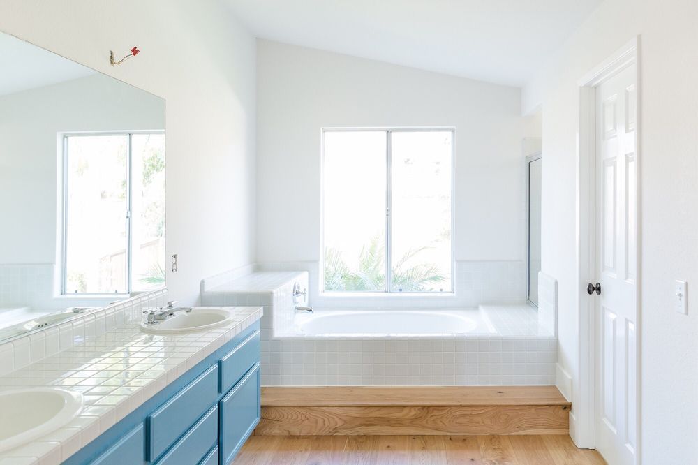 Bright white bathroom with a blue vanity, large window, and built-in bathtub on a platform.