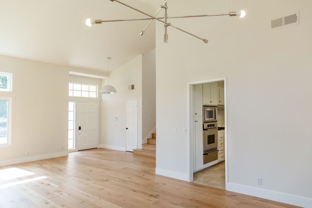 Spacious living room with light wood floors, white walls, and a modern chandelier. Entry to kitchen and stairs visible.