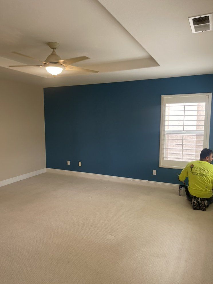 Bedroom with blue accent wall, beige walls/carpet, person kneeling by window, ceiling fan.