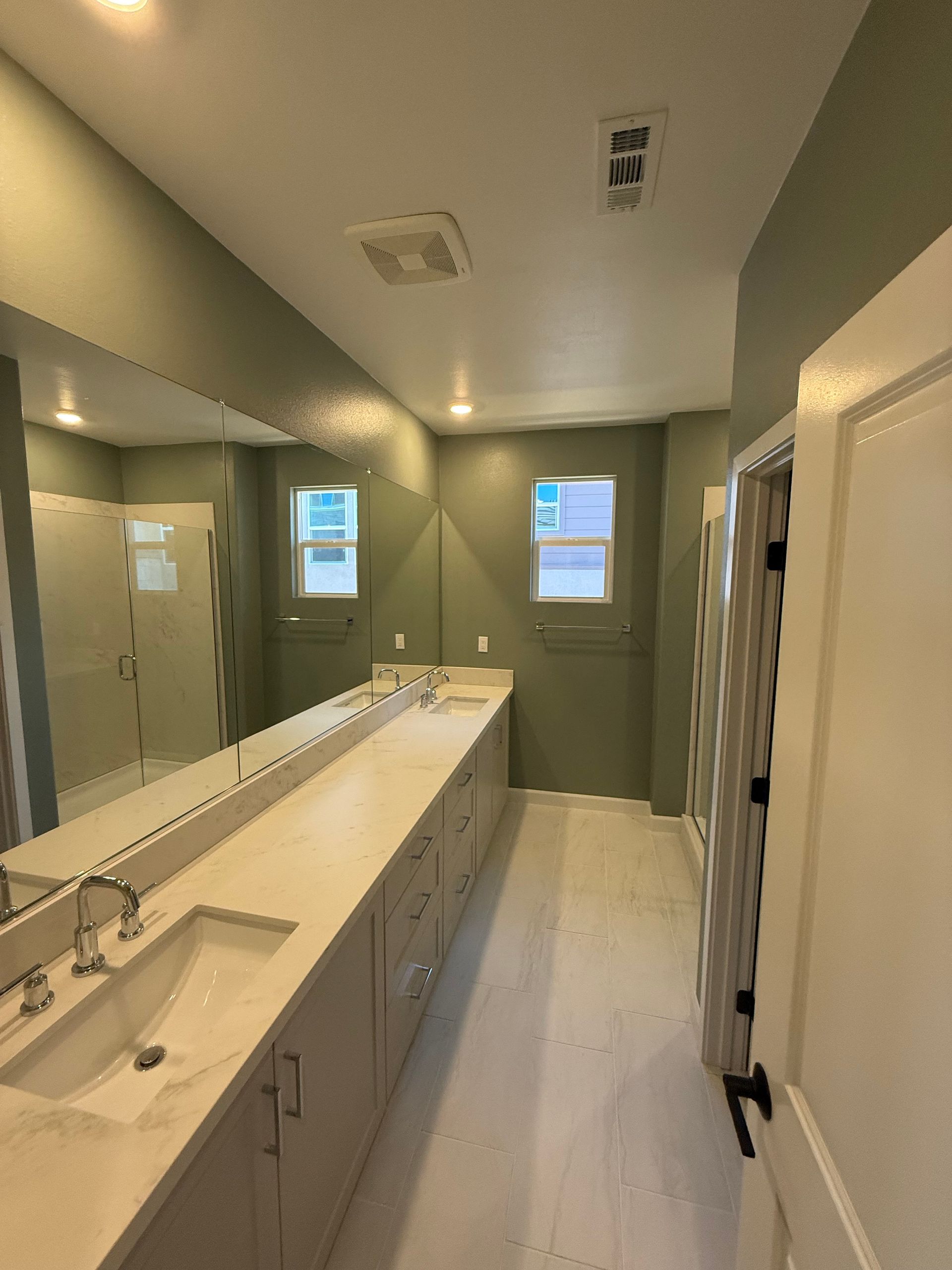 Spacious bathroom with long vanity, light cabinets, two sinks, and large mirror, green walls, and white floor.