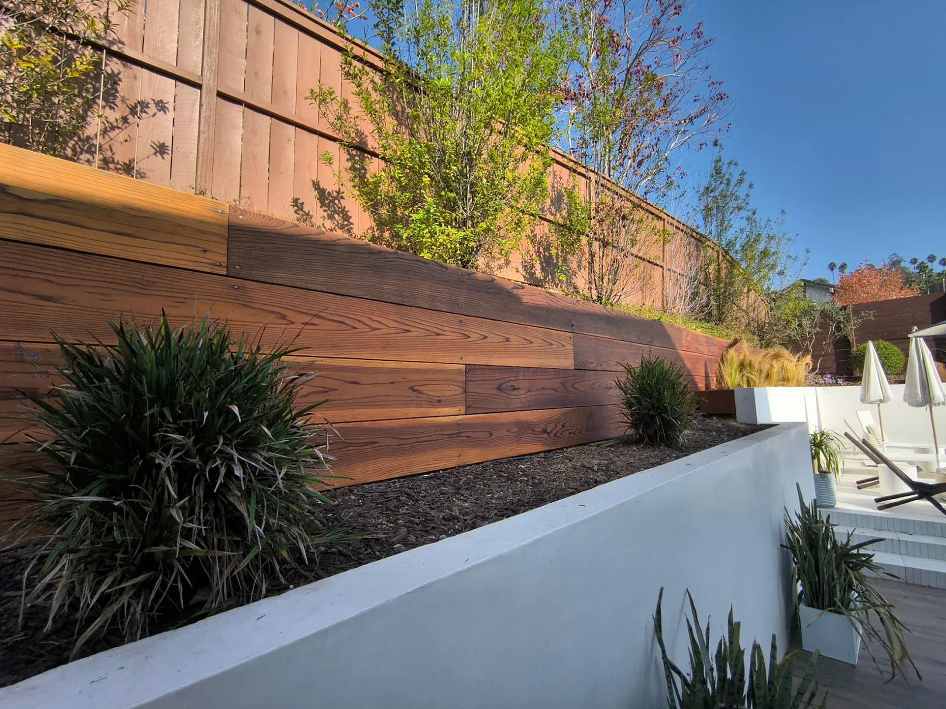 A wood retaining wall with plants and a fence, with a bright blue sky.