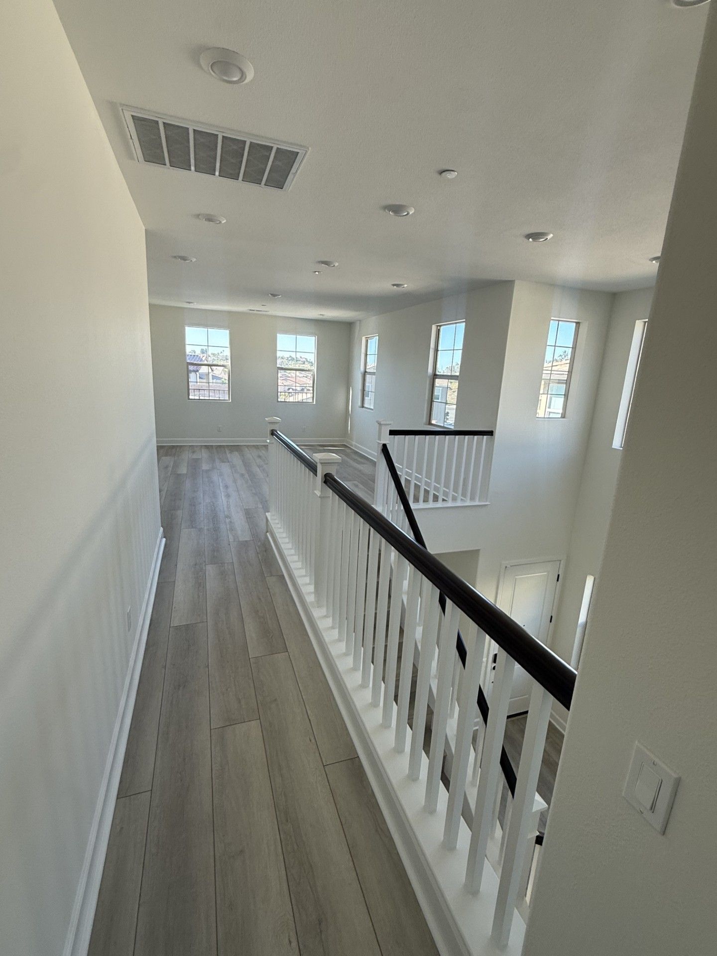 A bright, modern hallway with light-colored wood flooring, white walls, and a black-railed staircase overlooking a room.