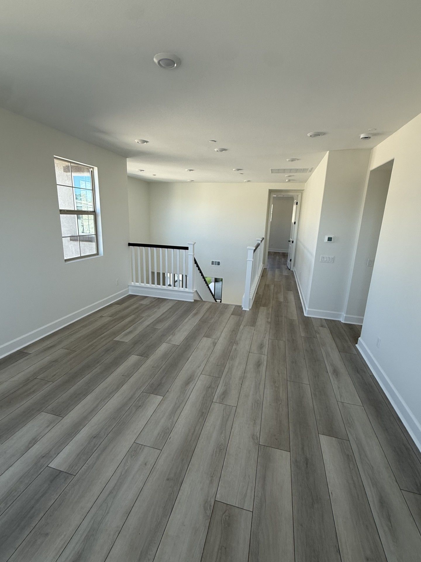 An empty room with light gray wood-look flooring, white walls, recessed ceiling lights, and a staircase landing.