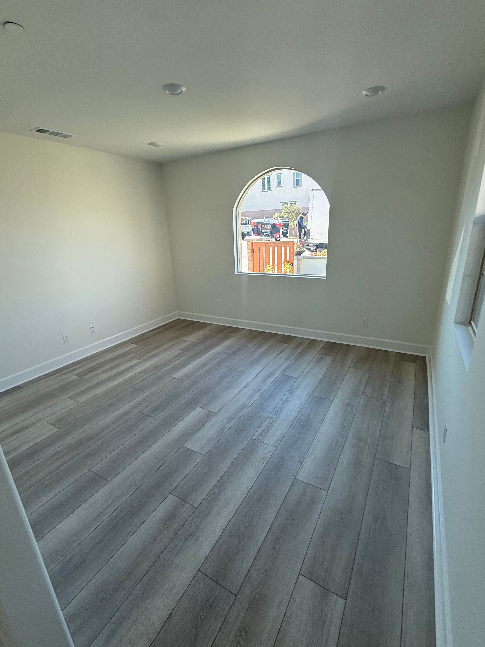 An empty, bright room with light gray wood-look flooring, cream-colored walls, recessed lighting, and an arched window.