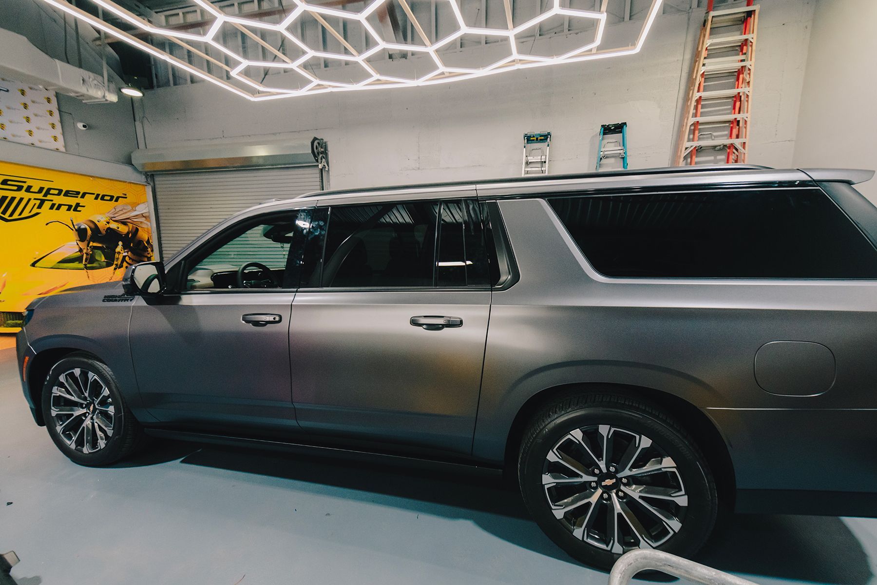 Grey SUV with tinted windows in a shop, under hexagonal lights, near a ladder and a yellow sign.