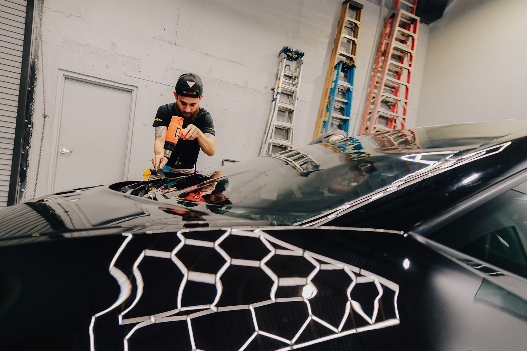 Man applying vinyl wrap to car with a honeycomb pattern; garage setting with ladders.