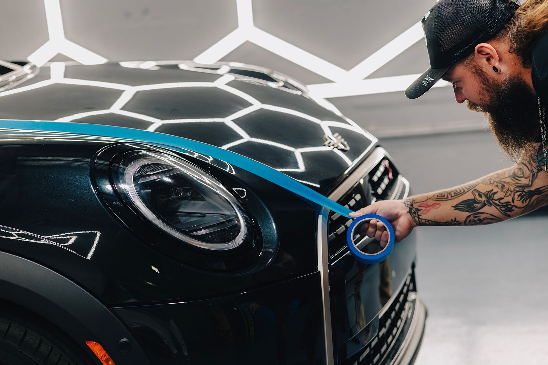 Person with beard taping a black car's front. Blue tape, shiny surface.