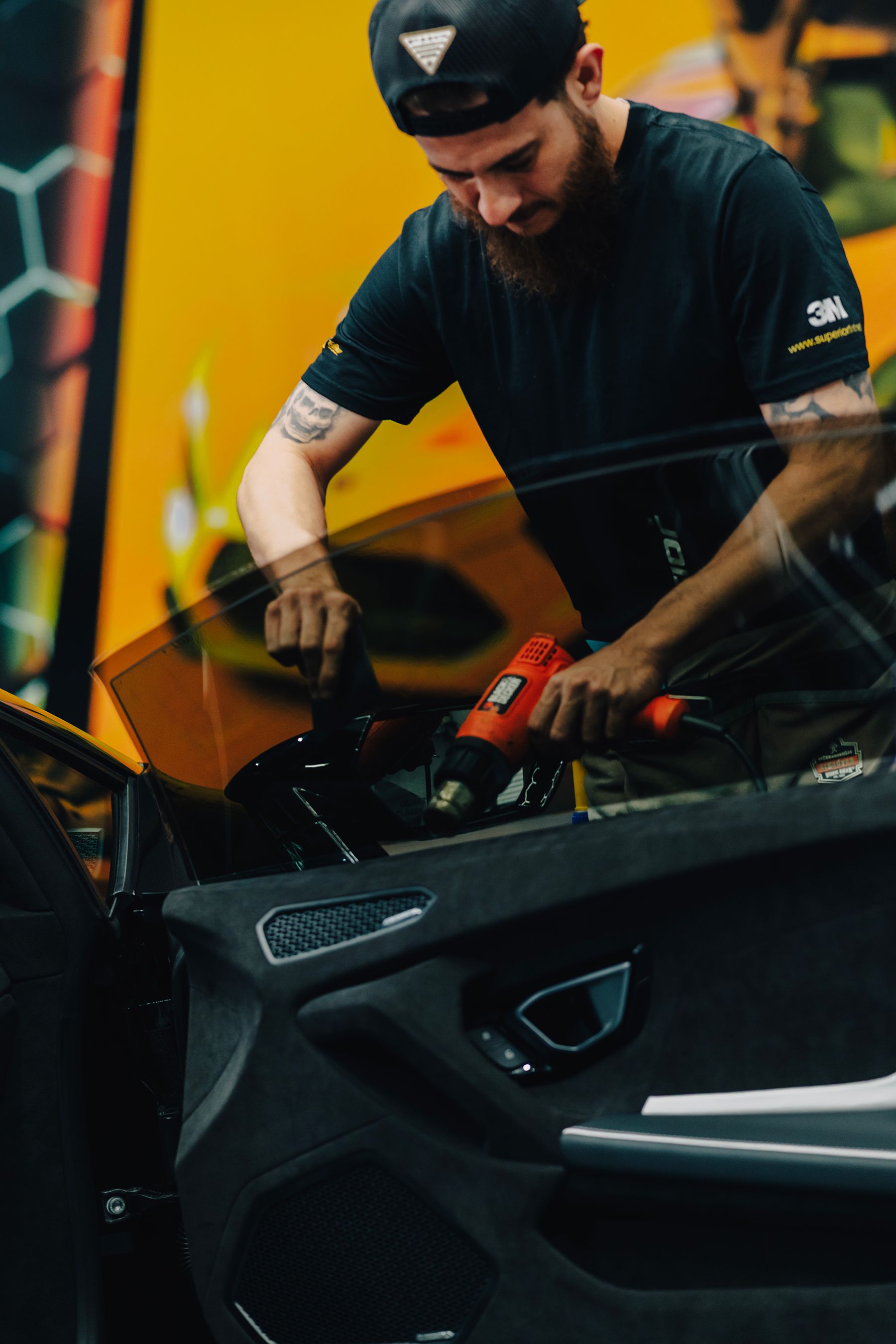Man applying tint to a car window with a heat gun. Indoors, focused, wearing a black cap and shirt.