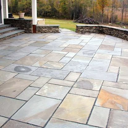 Stone patio with large gray pavers beside steps, a retaining wall, and a grassy yard with trees.