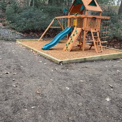 Wooden playground set with blue slide on a wood-chip pad beside a wooded area