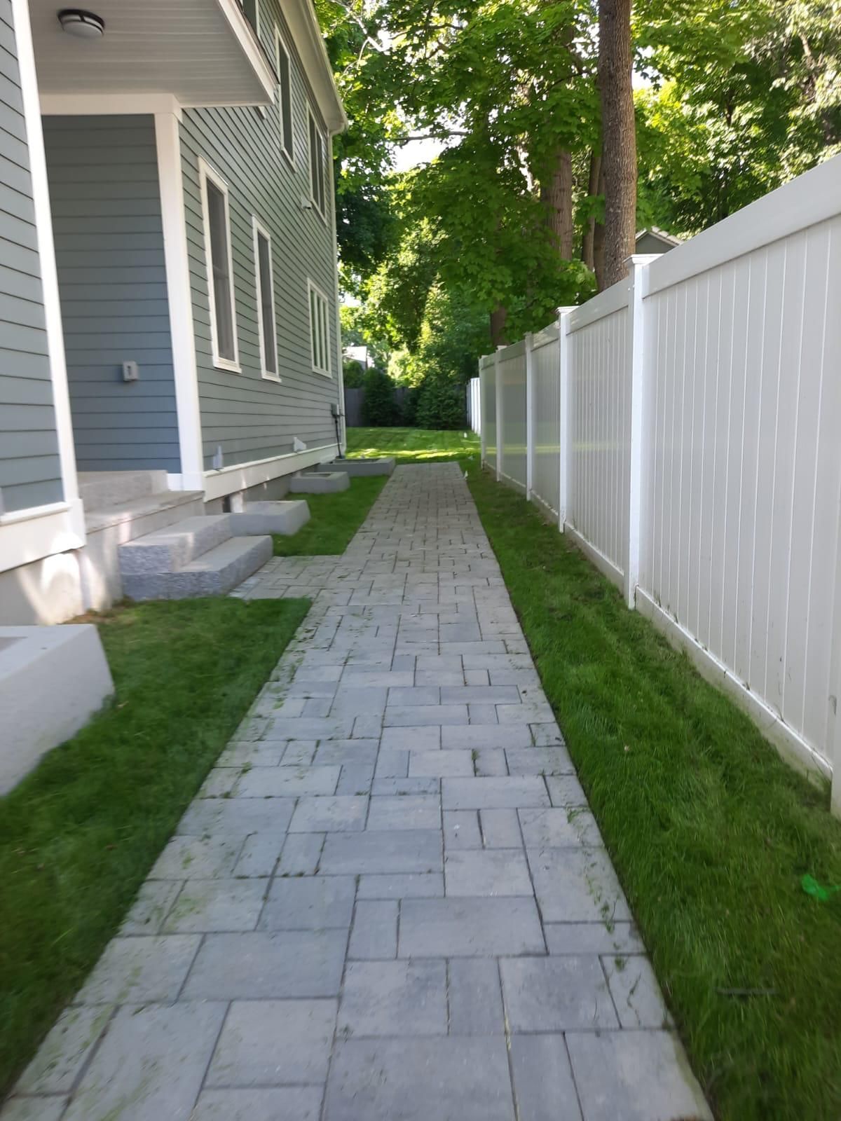 Narrow stone walkway beside a gray house and white fence, lined with grass and trees.