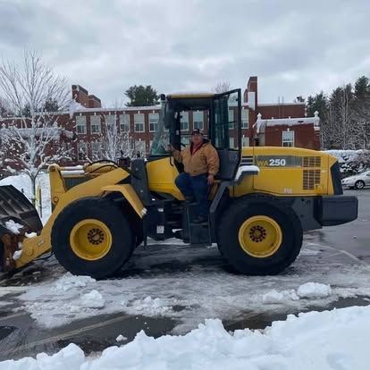 Yellow front loader with a person seated inside, parked on a snowy street near brick buildings.