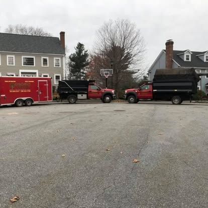 Red fire truck, dump truck, and pickup trucks lined up on a suburban street beside houses.