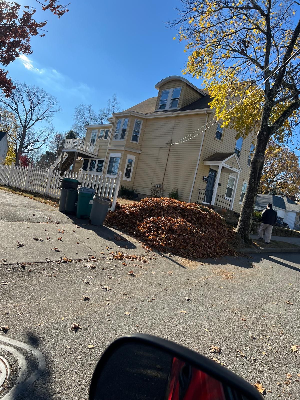 Tilted street view of a beige house with a leaf pile, trash bins, and a blue sky on a sunny day